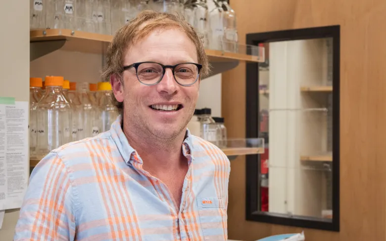 A smiling man in front of a shelf holding glass vials and a window door.