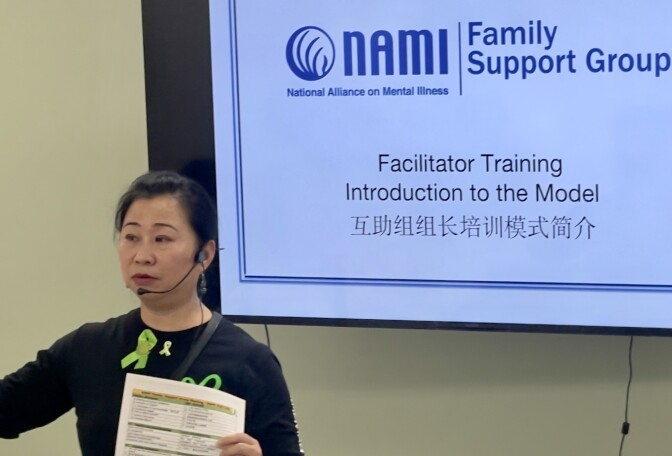 An Asian woman in a black shirt stands in front of a projector screen reading "Facilitator Training: Introduction to the Model."