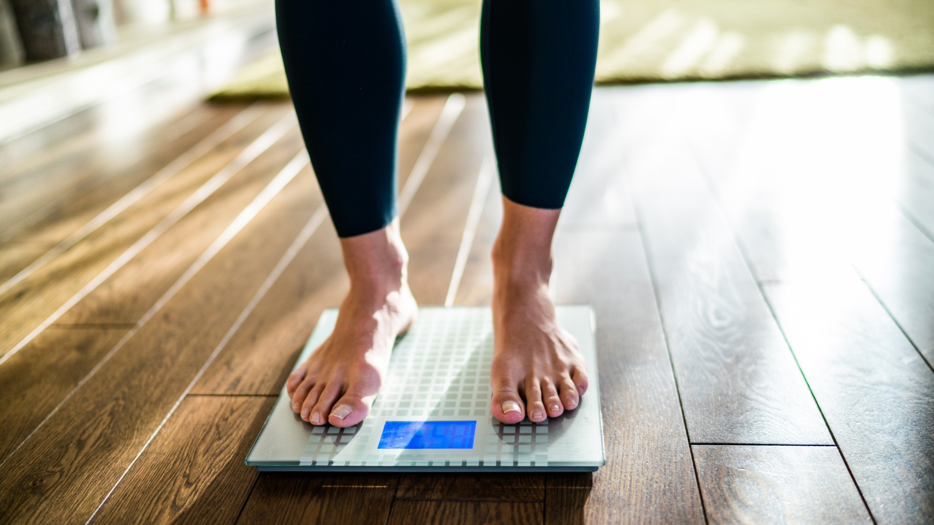 A woman's legs are shown standing on scales placed on a wooden floor. She wore black leggings and nothing on her legs.