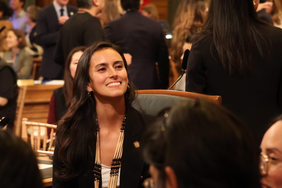 A woman sitting in a conference or meeting, surrounded by other attendees, smiles as she looks forward.