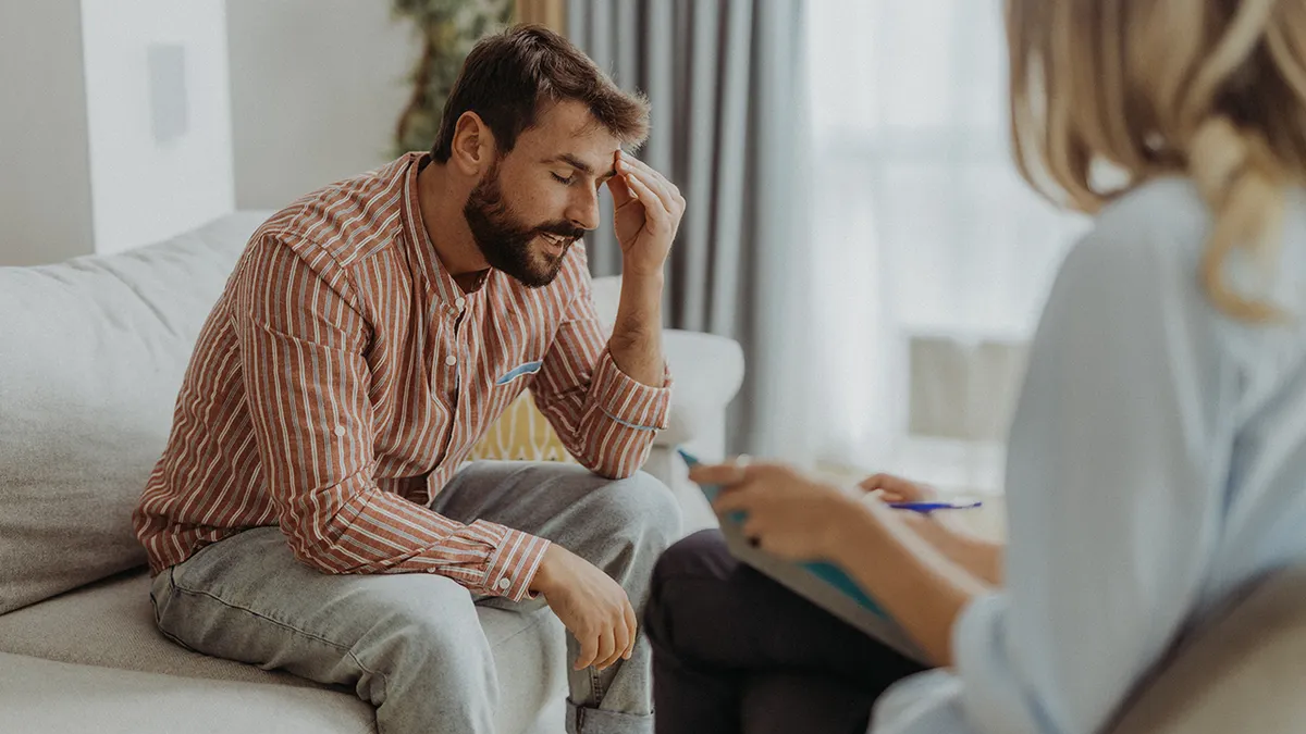 the man sits on the couch with his hand on his forehead while talking to the female therapist