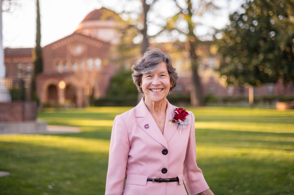 A woman wearing a light pink blazer with a red boutonniere stands smiling on a grassy campus lawn. Trees and a brick building with arched windows and a domed roof appear softly blurred in the background in warm afternoon light.