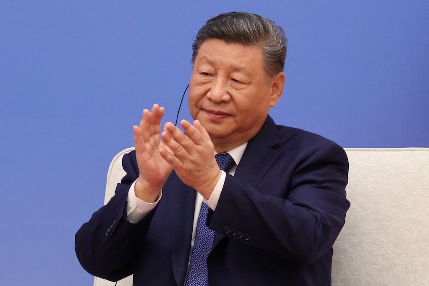 Sitting in a dark blue suit, Xi Jinping applauds.