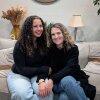 A young girl with curly brown hair sits on the bed next to her mother with shoulder-length, curly blonde hair. Both are wearing black sweaters with blue jeans. 