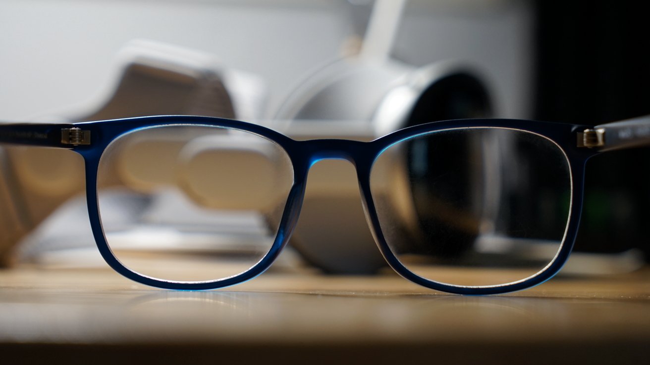Blue rectangular glasses resting on a wooden surface, in sharp focus, with dim white headphones and other desk items.