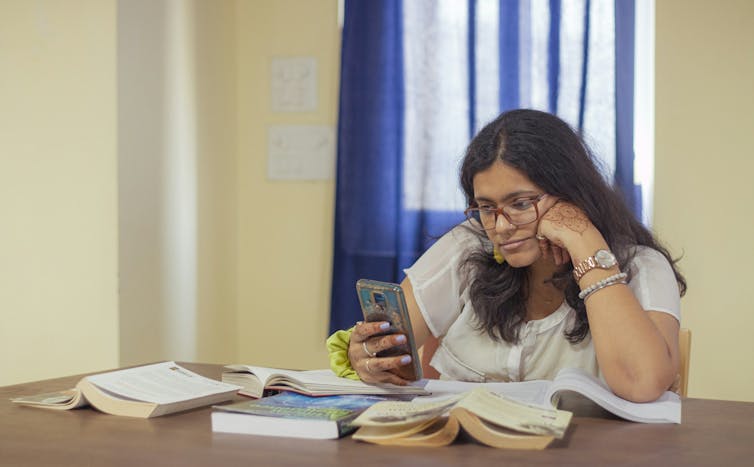 A woman with long hair and glasses stares at her phone surrounded by books at a table to study